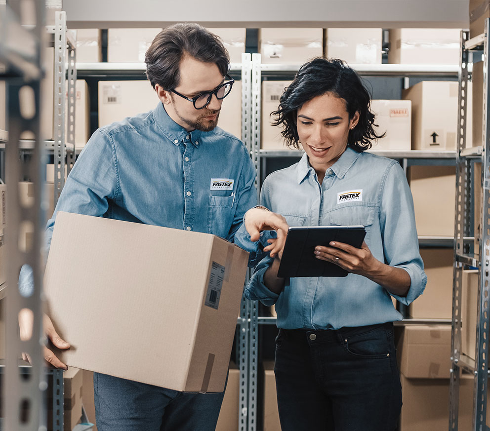 Two Fastex employees viewing a tablet together while in a warehouse holding a package.